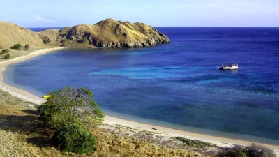 Vue panoramique d'une baie immaculée, probablement Pink Beach, dans le parc national de Komodo, avec des collines sèches et dorées, une eau bleu-turquoise cristalline et un bateau touristique ancré.
