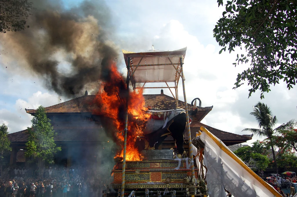 Large Balinese "cremation" ceremony in progress, with a tiered funeral pyre structure engulfed in flames and dark smoke, surrounded by crowds and a traditional building.