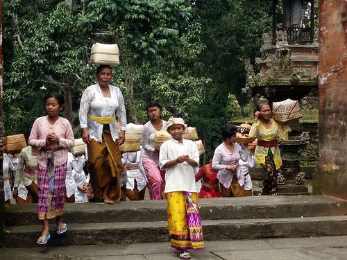 A group of Balinese women and a boy in traditional dress stand near a temple gate, carrying offerings on their heads during the "galungan" celebration.