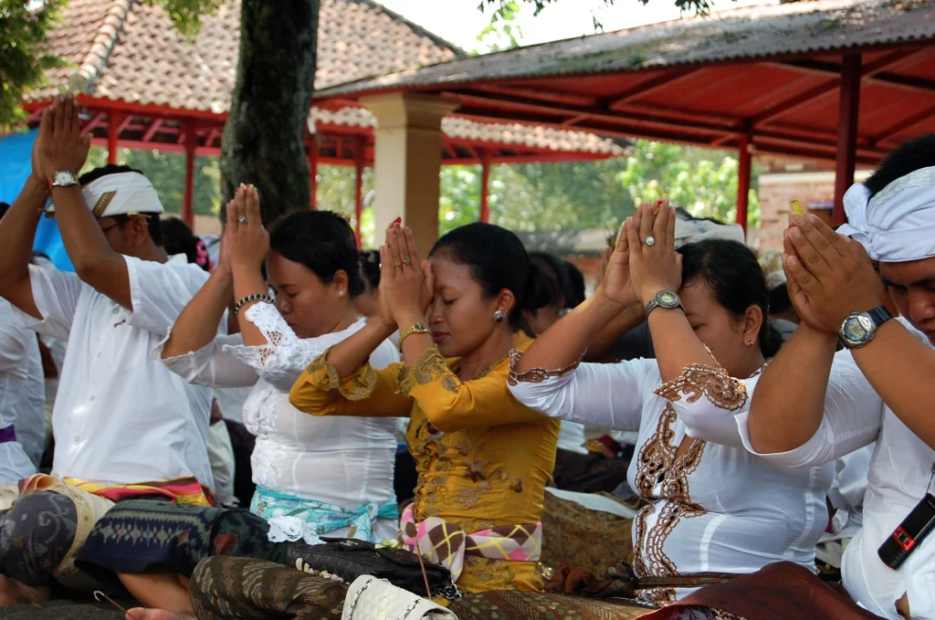 Balinese people sit and pray with hands pressed together at chest level in a communal temple setting for the "kuningan" holiday.