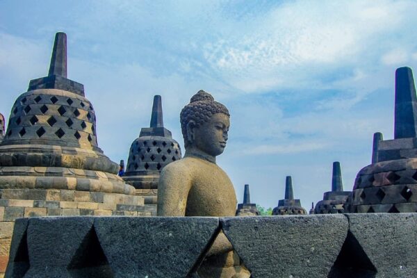 A stone Buddha statue on the upper terraces of the ancient "Borobudur" temple complex, surrounded by bell-shaped stupas under a cloudy sky.