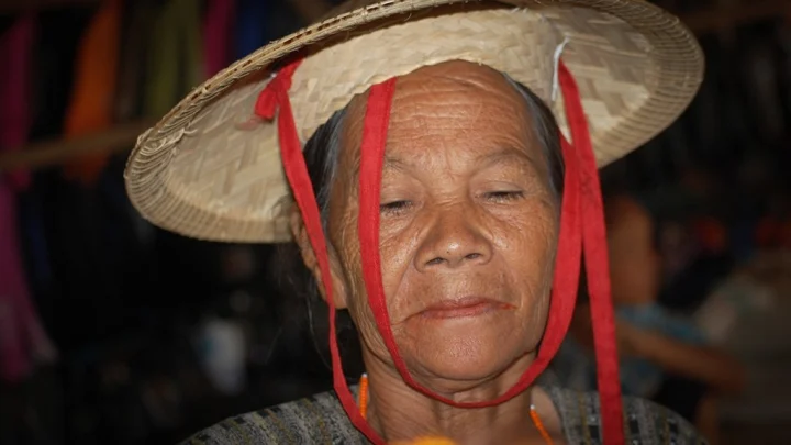 Close-up portrait of an elderly woman from Sulawesi wearing a woven conical hat with red ribbon chin straps.