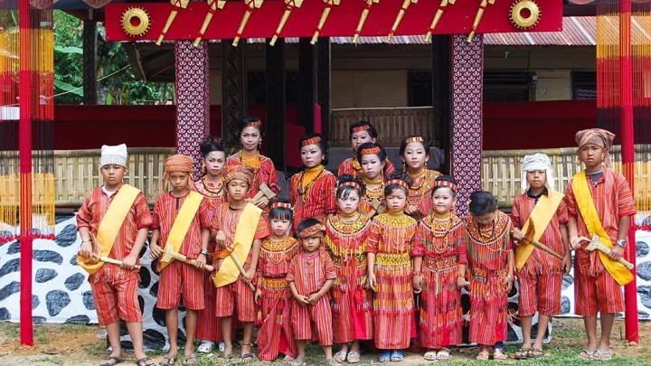 A group of children and women from Tana Toraja, Sulawesi, wearing bright red and orange striped traditional garments, posing in front of an ornate traditional house