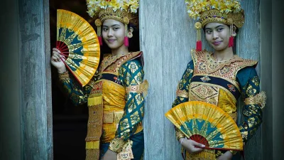 Portrait de deux jeunes danseuses balinaises en costumes traditionnels élaborés, coiffes dorées ornées de fleurs et tenant des éventails jaunes et rouges, prêtes à exécuter une danse.